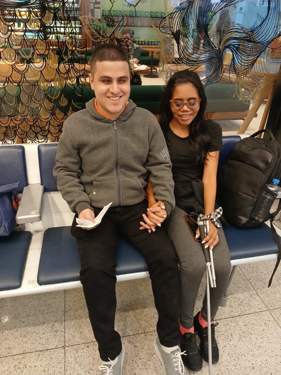 Casal sentado em um banco de aeroporto. O homem está à esquerda, sorrindo, usando uma jaqueta cinza, calça preta e tênis cinza. Ele segura um papel na mão esquerda e está de mãos dadas com a mulher ao lado. A mulher está à direita, usa óculos de lentes azuladas, camiseta preta, calça cinza escura, meias rosa e sapatilhas pretas. Ela segura uma bengala branca, sinalizando deficiência visual. Ambos parecem felizes e relaxados. Ao lado da mulher há uma mochila preta. No fundo, há uma divisória de vidro decorada com desenhos pretos e dourados.