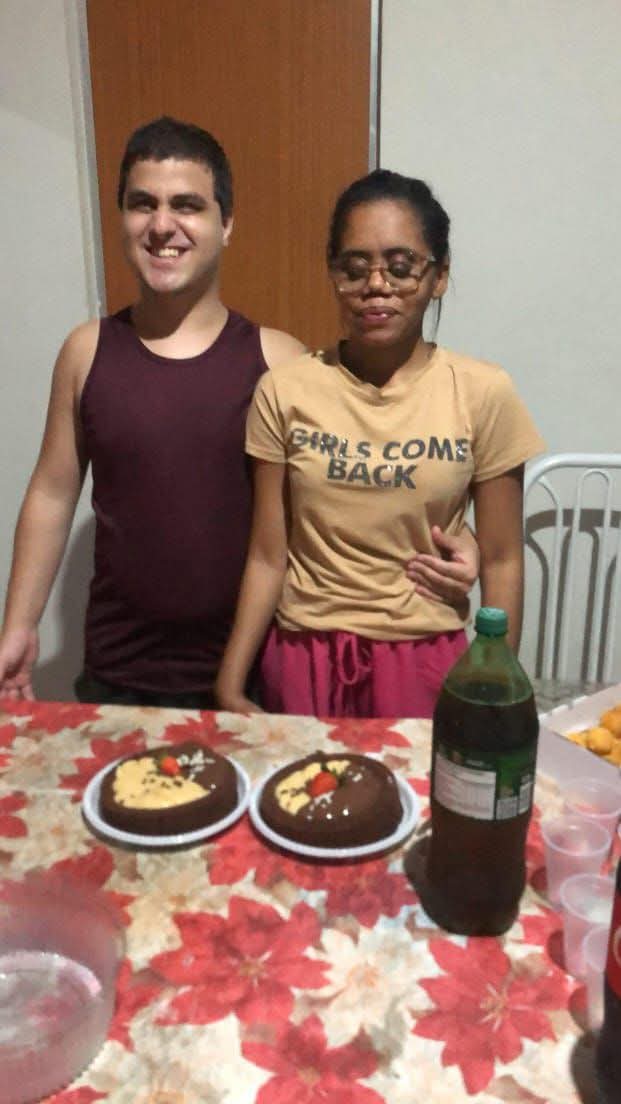 Foto de duas pessoas em pé, próximas uma da outra, sorrindo, atrás de uma mesa decorada com toalha de flores vermelhas e brancas. A pessoa à esquerda é um homem com cabelo curto castanho escuro, vestindo uma camiseta regata marrom escura. Sua mão está apoiada no ombro da pessoa ao lado. A pessoa à direita é uma mulher de pele mais escura, óculos, cabelo preso, usando uma camiseta amarela com os dizeres "GIRLS COME BACK" e shorts rosa. Ela mantém os olhos fechados e uma expressão tranquila. Sobre a mesa estão dois bolos redondos cobertos com chocolate, decorados com pedaços de chocolate branco e morangos inteiros em cima. Também há uma garrafa de refrigerante verde (provavelmente guaraná), alguns copos plásticos transparentes e uma tigela de plástico vazia no canto esquerdo inferior da imagem. Ao fundo, aparecem uma cadeira branca de metal e uma porta de madeira.