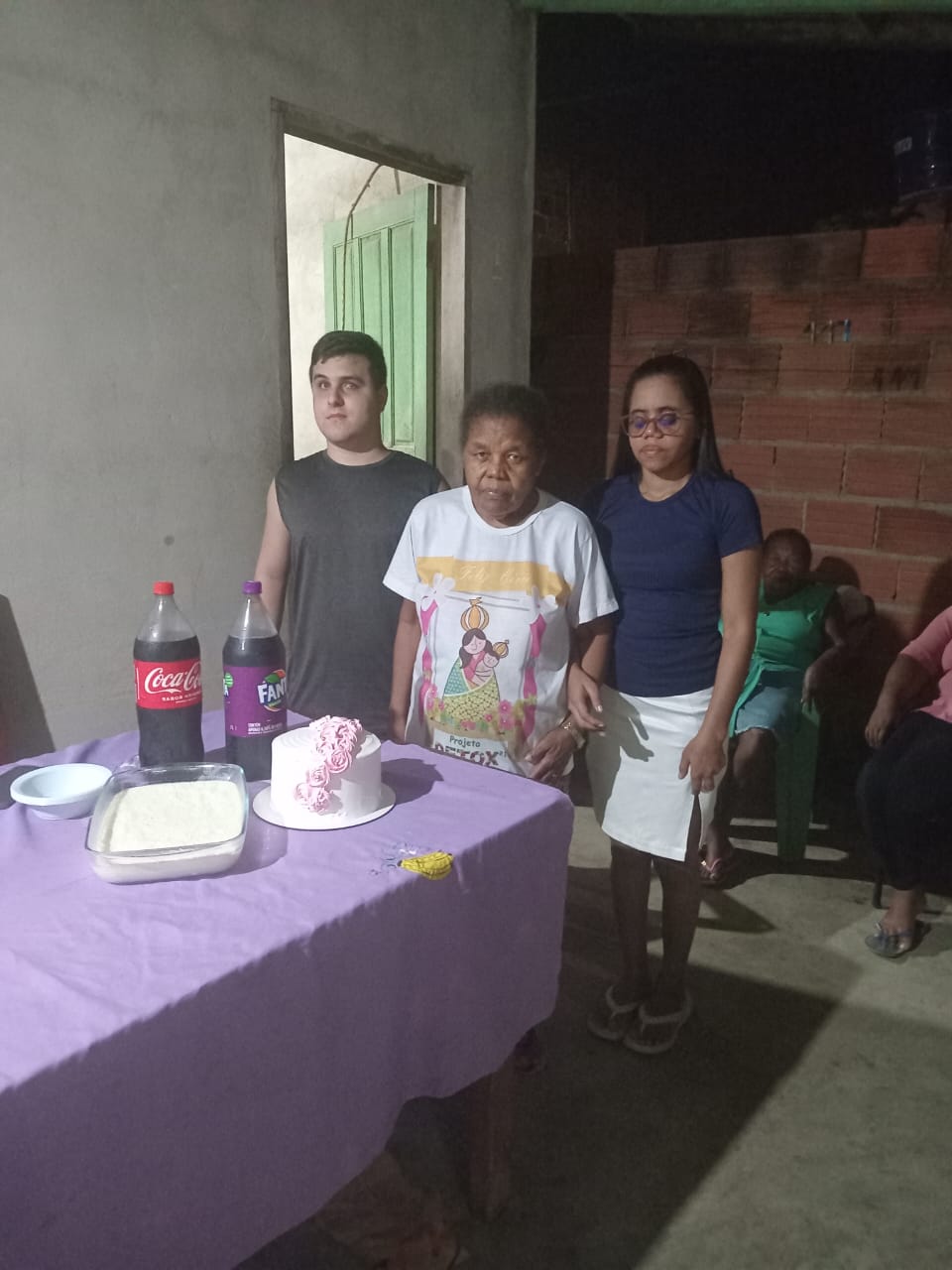 Três pessoas em pé, posando para foto em uma comemoração simples dentro de uma casa. À frente delas, há uma mesa coberta com uma toalha roxa, nela há dois refrigerantes (Coca-Cola e Fanta), um bolo branco decorado com rosas cor-de-rosa, uma travessa de comida e um prato branco vazio. A mulher ao centro veste uma camiseta branca estampada, enquanto a pessoa à esquerda (um homem jovem) está com uma camiseta preta sem mangas. A pessoa à direita (uma jovem) está com uma blusa azul escura e saia branca, além de usar óculos. Ao fundo, há pessoas sentadas em cadeiras plásticas e uma parede de tijolos aparentes. O ambiente é simples, com luz artificial e clima de confraternização.