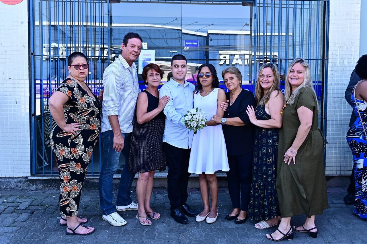 Foto de grupo em frente a um prédio com portas de vidro. O centro da imagem é ocupado por um casal, aparentando serem noivos (a mulher veste um vestido branco e segura um buquê de flores, enquanto o homem usa camisa clara). Ambos estão cercados por familiares e amigos, formando uma fileira. As pessoas sorriem e demonstram alegria, algumas tocando ou abraçando os noivos. O clima é descontraído e feliz, típico de celebração de casamento civil. Ao fundo, vê-se parte da fachada do prédio, grades e sinais.