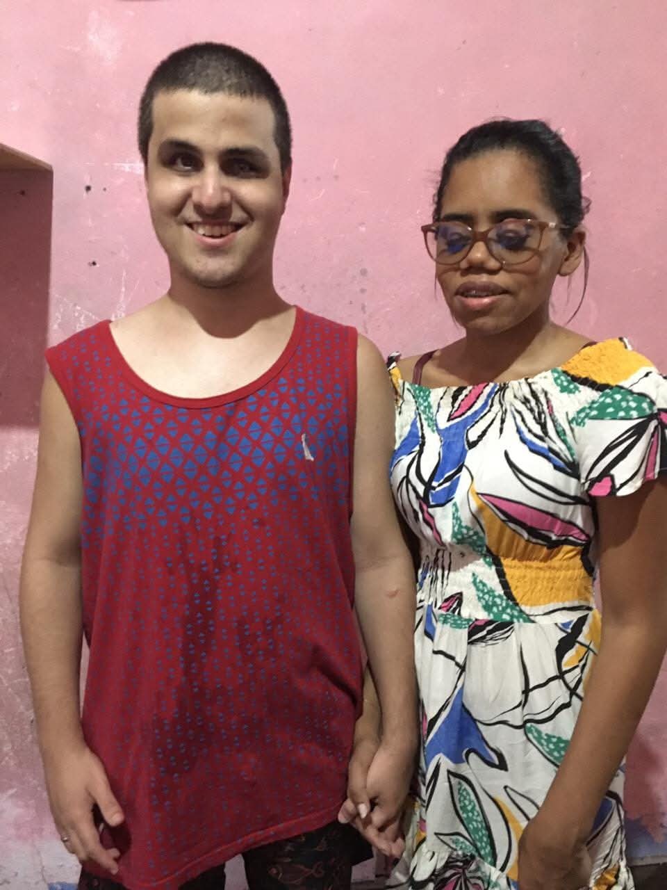 Duas pessoas de pé, de mãos dadas, em frente a uma parede rosa. À esquerda está um homem jovem de cabelos curtos, pele clara, usando uma regata vermelha com detalhes em azul e sorri para a câmera. À direita, uma mulher jovem de pele escura, usando óculos de grau e um vestido branco estampado com cores vivas, cabelo preso. Ela está com os olhos fechados e também sorri levemente. O ambiente parece simples e acolhedor.