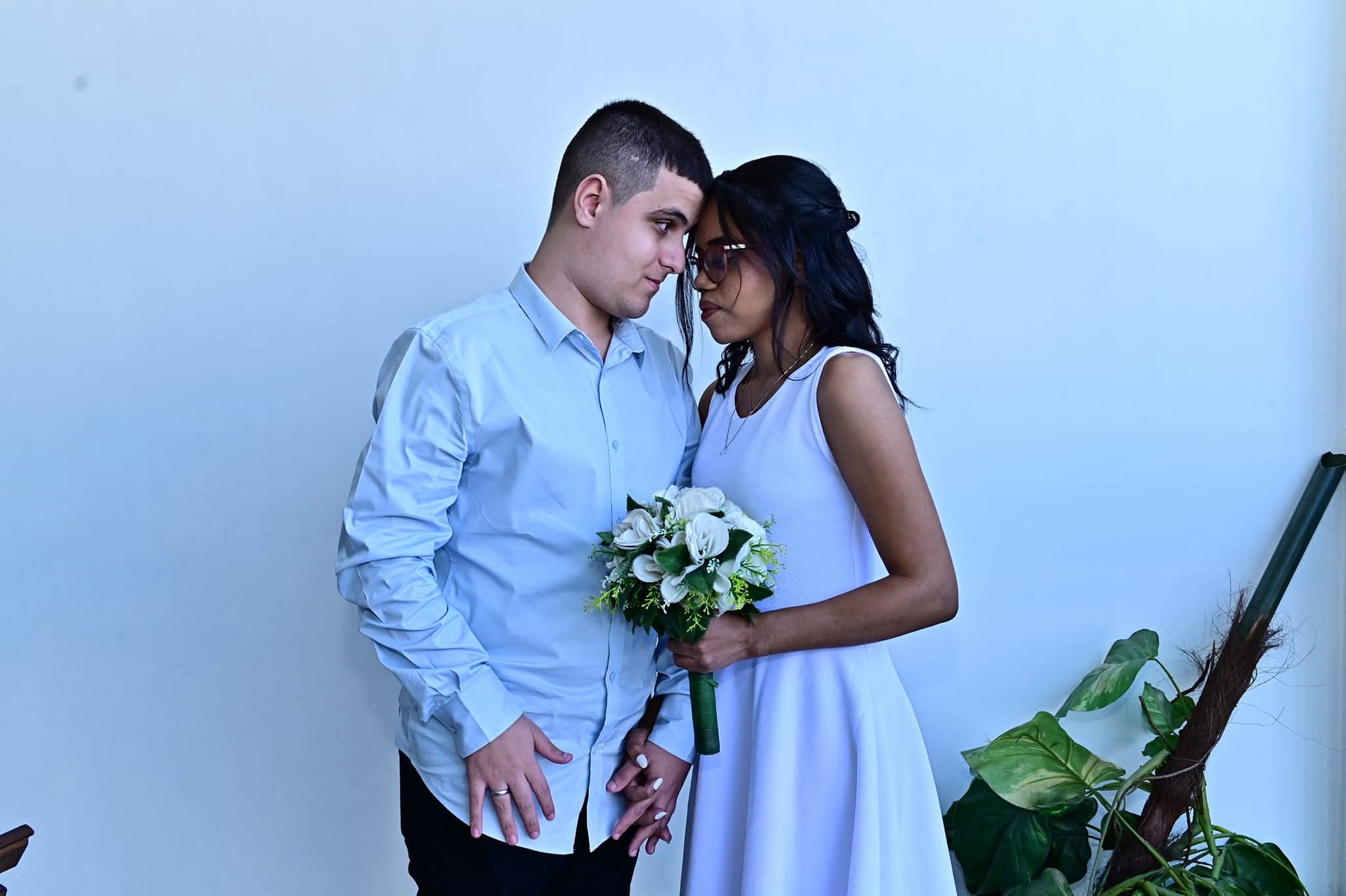 Casal jovem posando para uma foto, com um fundo branco e algumas plantas ao lado direito. Eles estão de frente um para o outro, encostando as testas em um gesto de carinho. O homem veste uma camisa social azul clara e calça preta, e segura a mão da mulher. A mulher usa um vestido branco simples e segura um buquê de flores brancas com folhas verdes, além de usar óculos e ter cabelos escuros e soltos. O clima parece romântico e íntimo, sugerindo que a ocasião pode ser especial, como um casamento civil