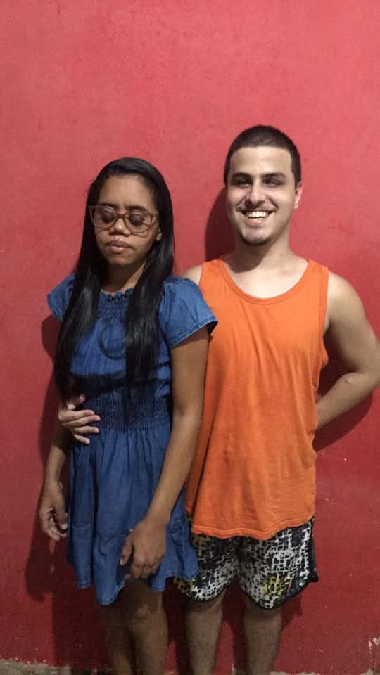 Duas pessoas em pé em frente a uma parede vermelha. À esquerda está uma mulher de cabelos longos e lisos, usando óculos grandes, vestido azul e expressão séria ou triste, com os olhos fechados. À direita está um homem de cabelo curto, sorrindo, vestindo uma regata laranja e bermuda estampada em preto, branco e amarelo. Ele está com o braço esquerdo atrás das costas e segura com a outra mão a cintura da mulher.